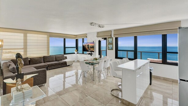 A modern living room with a sofa, dining table, and chairs, large windows with a view of the sea, and a flat-screen TV. - Stylish Oceanfront 2 BR 1.5 BA Condo with Resort Amenities, Pools, Gym, Sauna 1402 (Miami Beach)