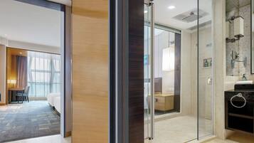 A hotel room with a glass-enclosed shower, a wooden floor, a desk with a chair, and a bed.