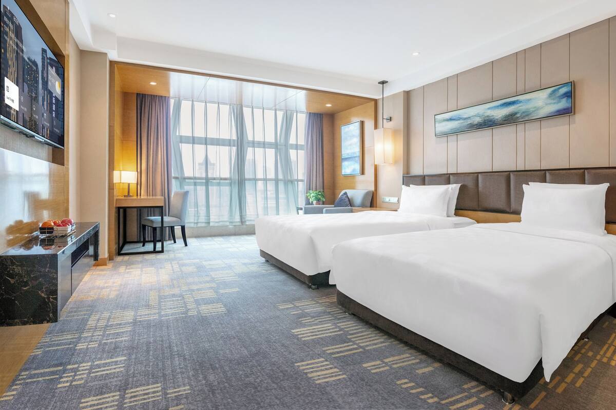 Habitación de hotel con dos camas, un escritorio, una silla, un ventanal grande y un cuadro en la pared.
