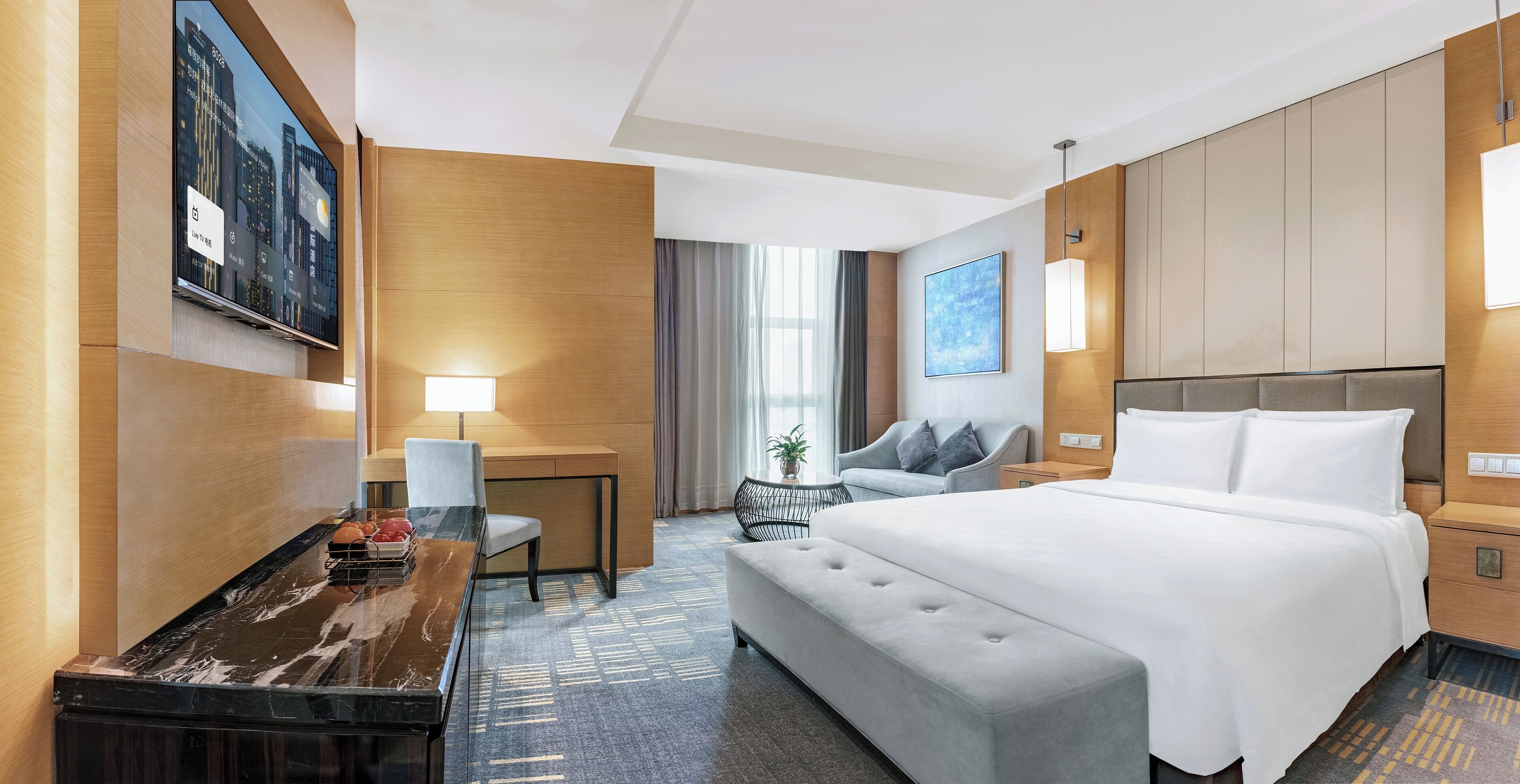 Een hotelkamer met een bed, een bureau, een stoel, een bankje, een lamp en een televisietoestel dat een stadsgezicht toont.