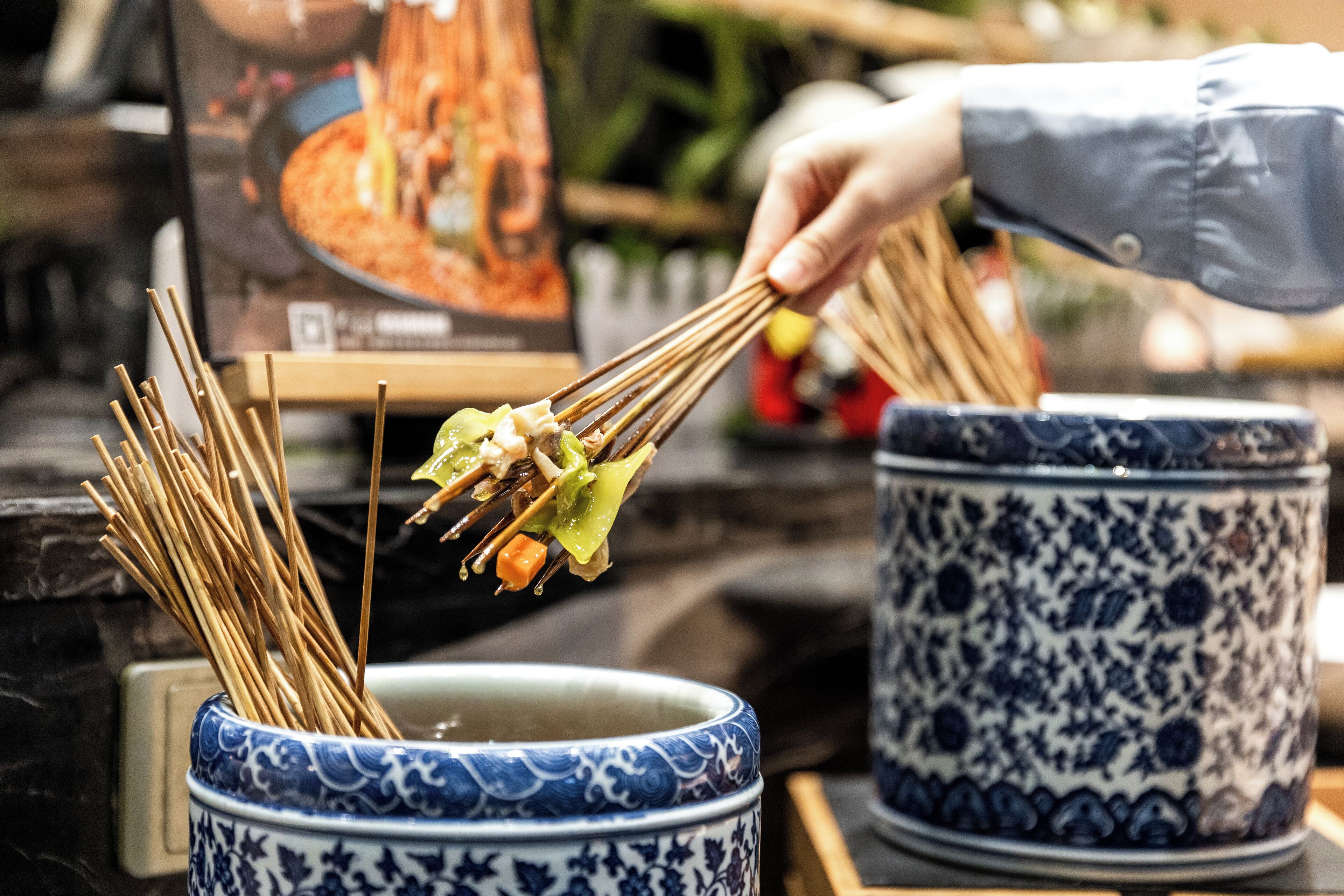 A hand holding chopsticks over a bowl of food, with a decorative blue and white ceramic container in the background.