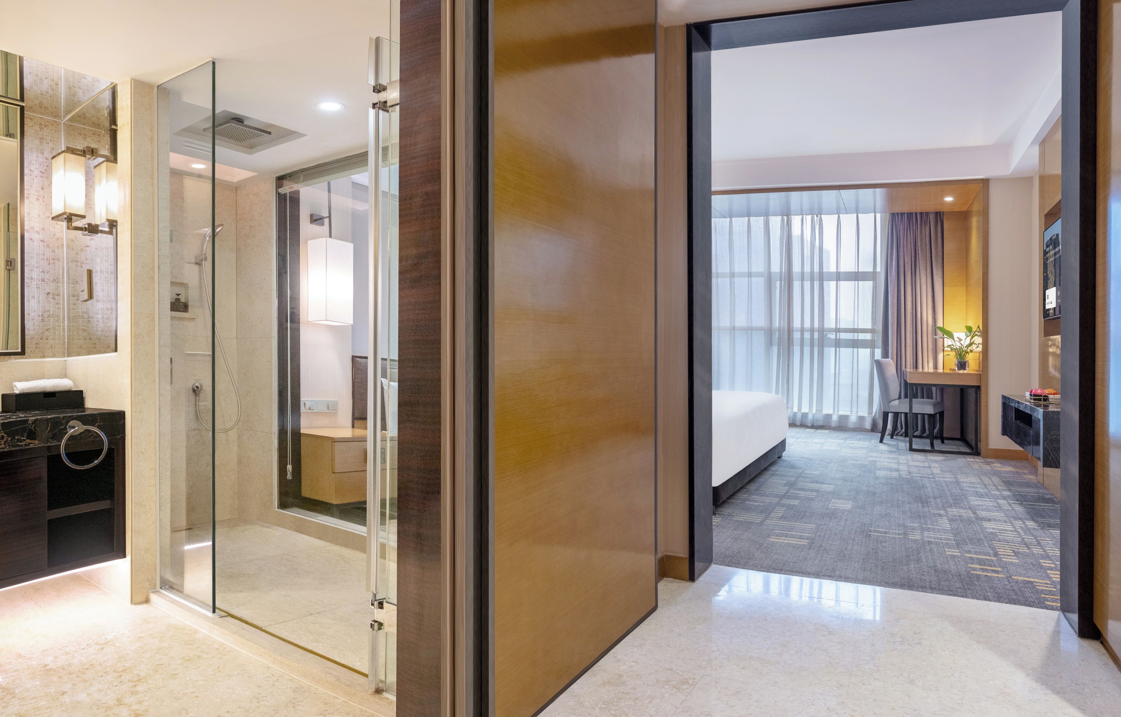 A hotel room with a glass-enclosed shower, a wooden vanity, a bed, a desk with a chair, and a large window with curtains.