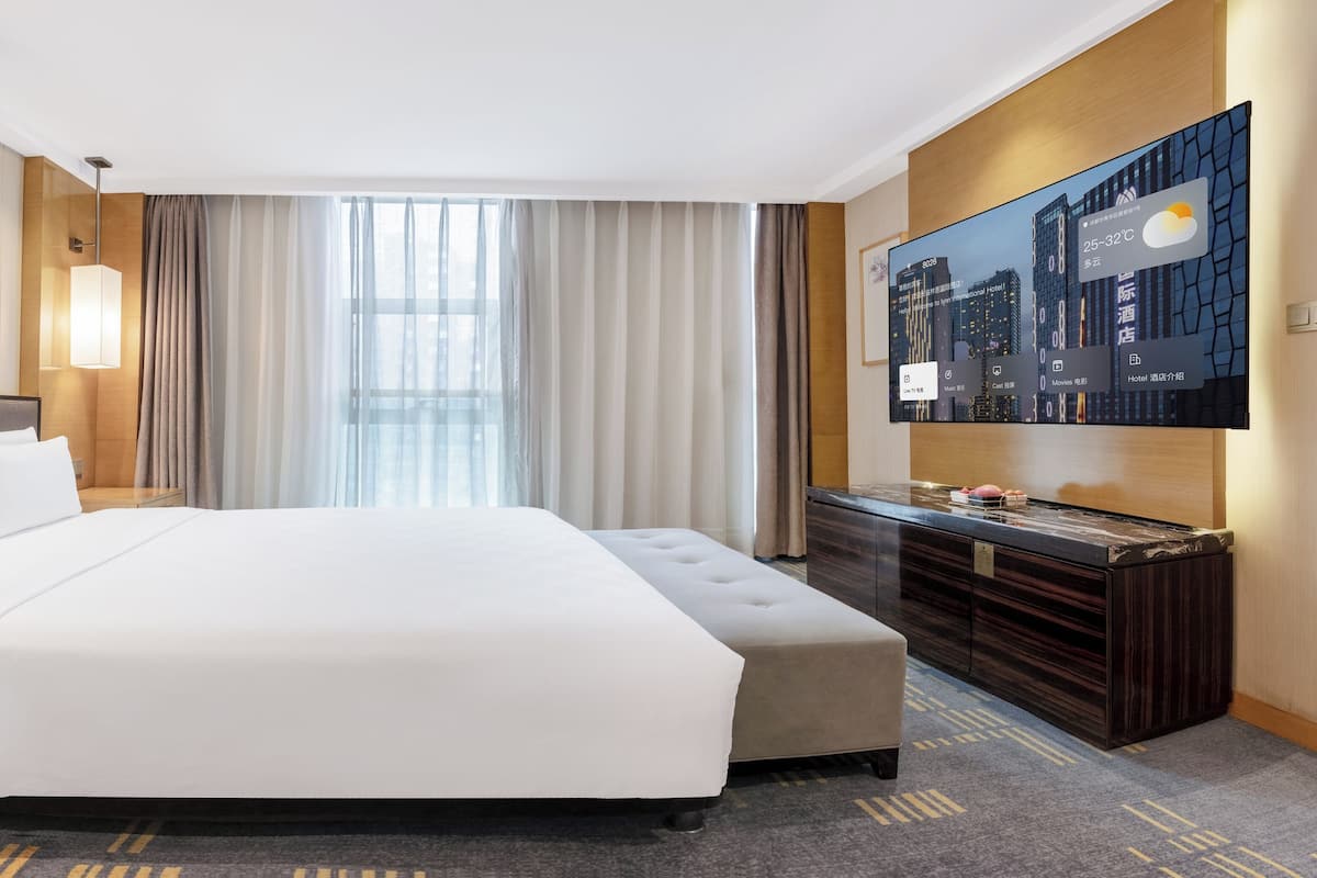 Habitación de hotel con una cama grande, un televisor de pantalla plana montado en la pared, un aparador de madera y un ventanal con cortinas ligeras.