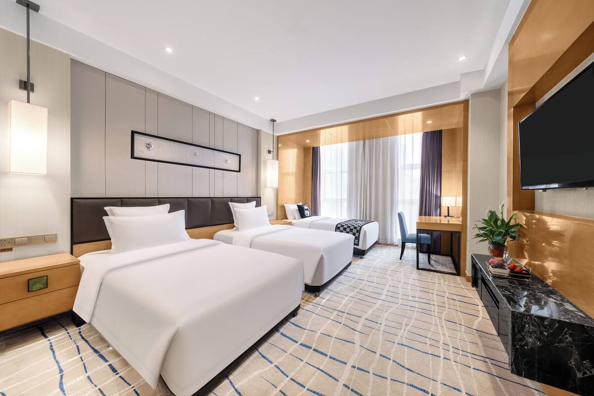 Habitación de hotel con dos camas, un televisor de pantalla plana, un escritorio con una silla y un ventanal con cortinas.