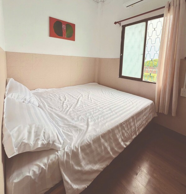 A single bed in a room with a plain wall, a window with curtains, and a small picture frame.