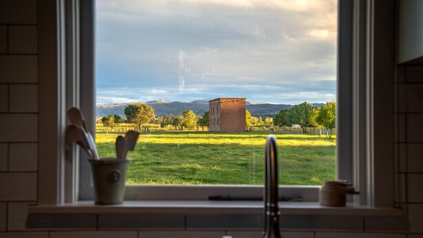 A kitchen with a window view of a grassy field, a brick building, and mountains in the distance. - Historic Castle Coach House – Trail & Wineries (Earnscleugh)