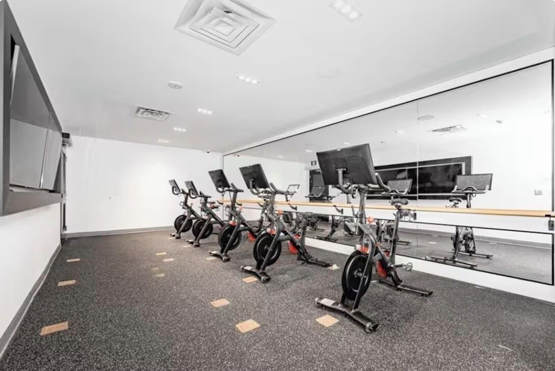 A gym with stationary bikes arranged in a row, a large mirror wall, and a television screen.