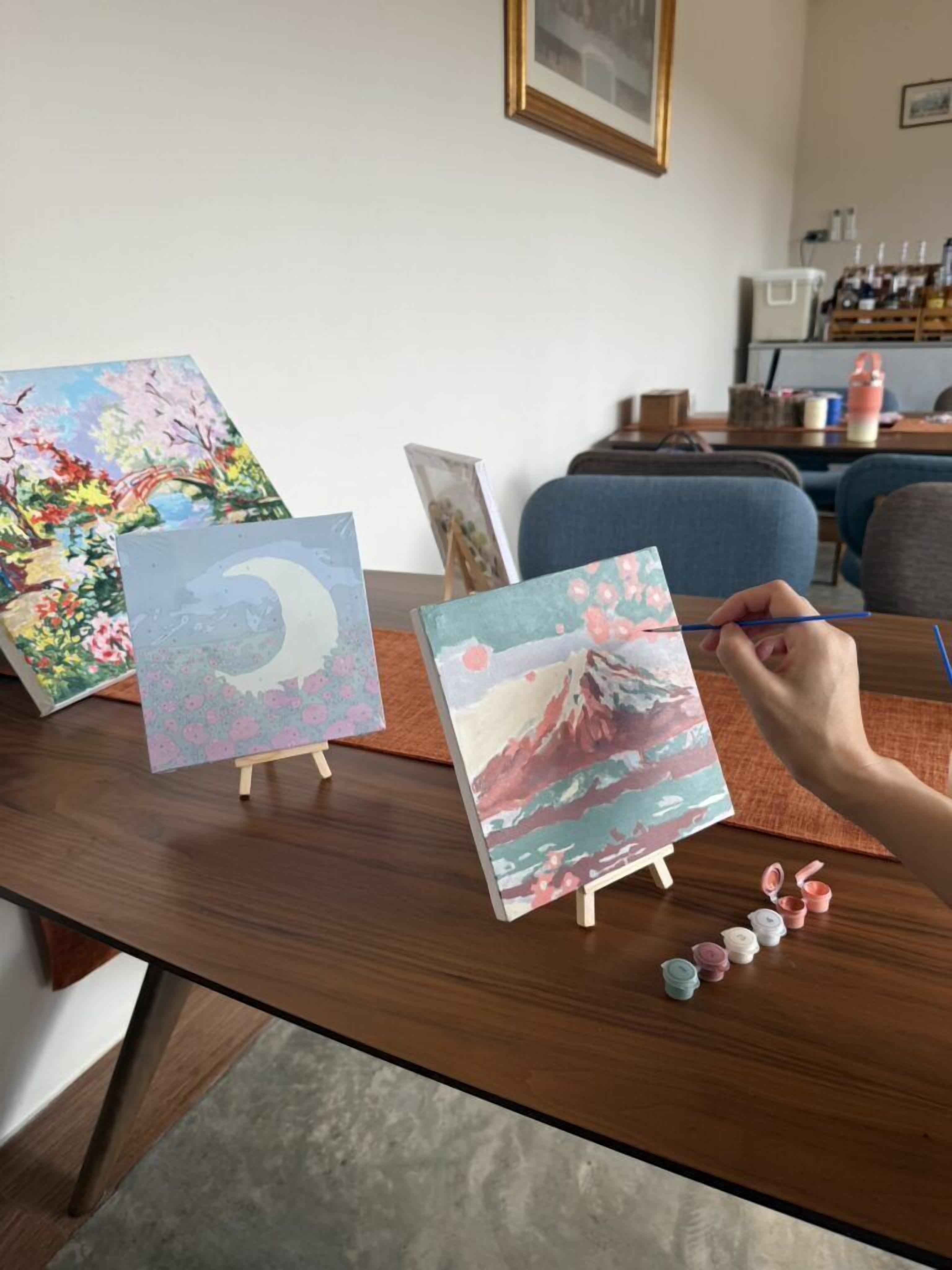 Un atelier de peinture avec des chevalets sur lesquels sont exposées des peintures colorées, une personne en train de peindre et du matériel d’art disposé sur une table en bois.