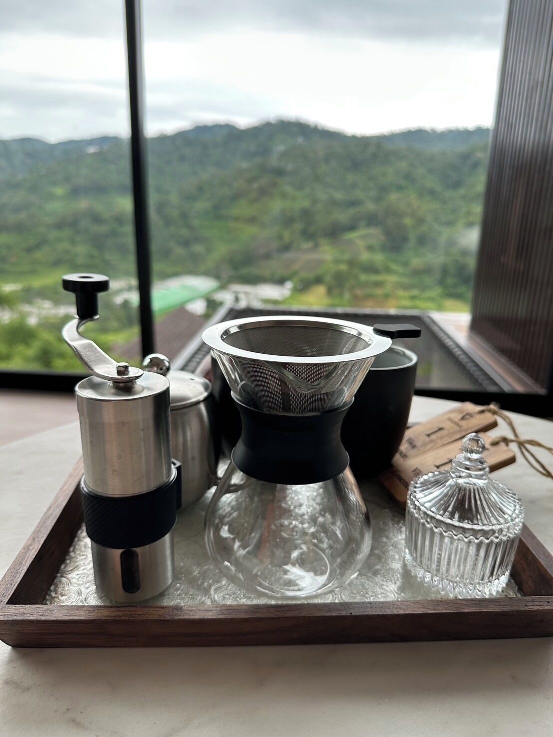 Un plateau contenant un moulin à café, un filtre à café et une carafe en verre, posé sur un rebord de fenêtre d’où l’on peut admirer des collines verdoyantes.