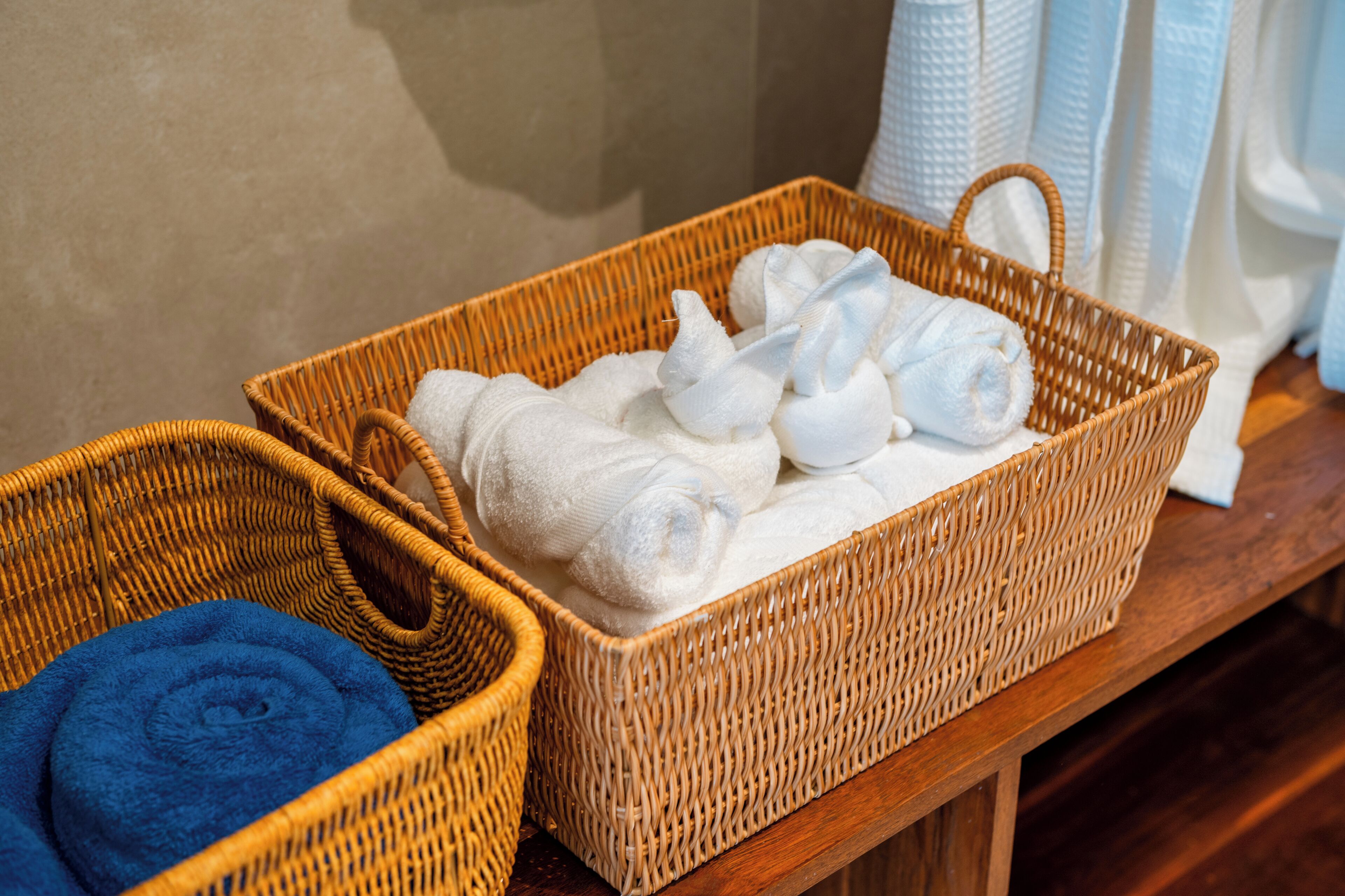 Deux paniers en osier, l’un contenant des serviettes blanches et l’autre une serviette bleue, sont posés sur une table en bois.