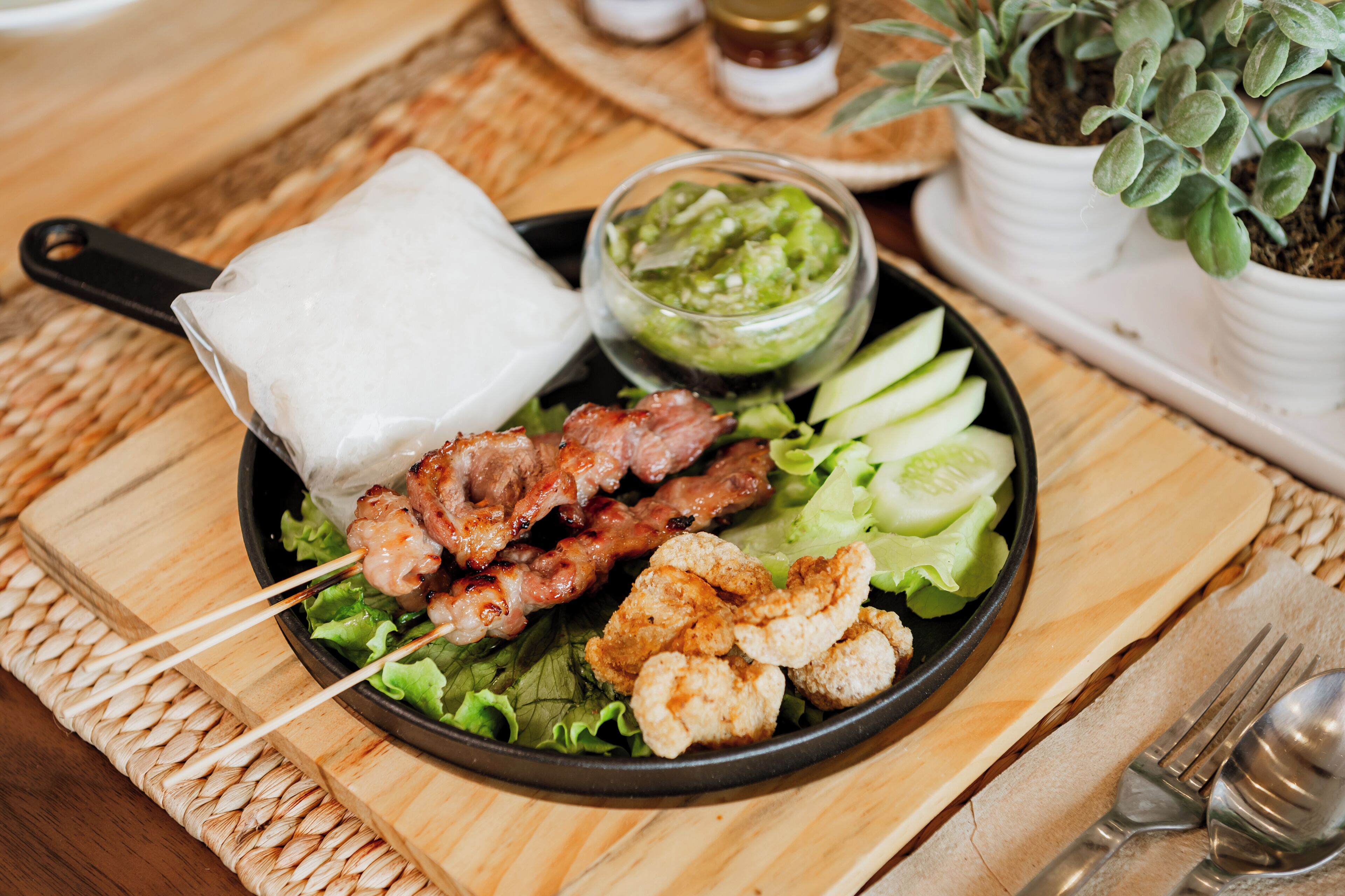 Un plato de brochetas de carne, un recipiente con salsa y rodajas de pepino sobre una mesa de madera.