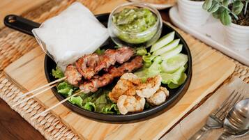 Un plato de brochetas de carne, un recipiente con salsa y rodajas de pepino sobre una mesa de madera.