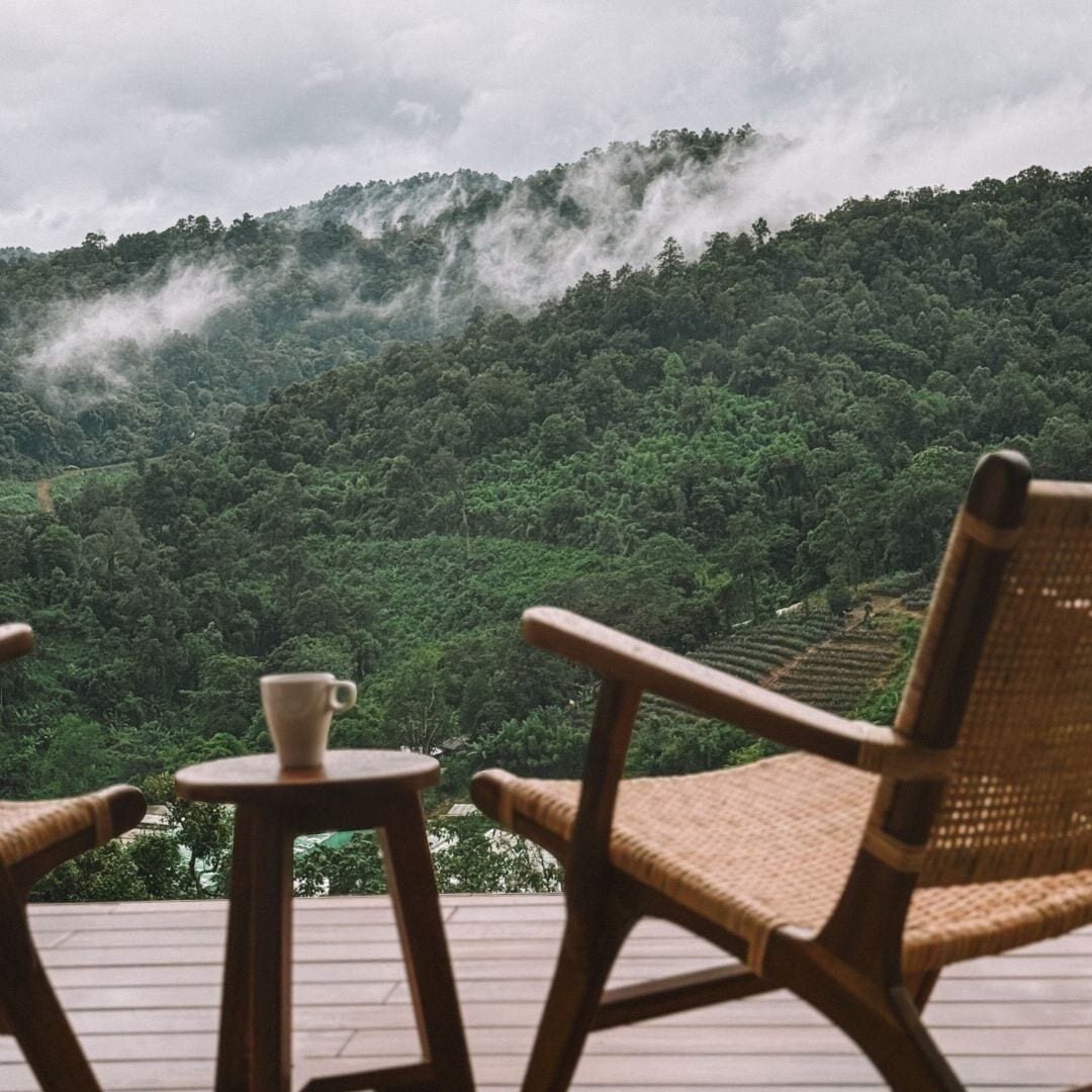 A wooden deck with wicker chairs and a small table, overlooking a lush green forest with mist rising from the hills.