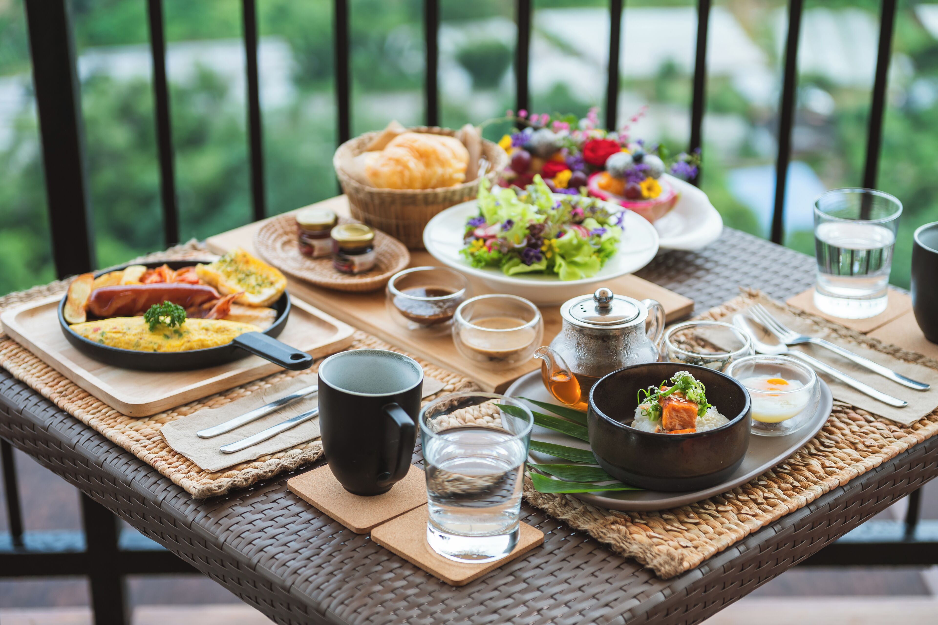 A breakfast setup on a wicker table with various dishes, including eggs, toast, and a salad, along with cups and a teapot.