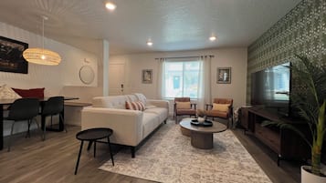Un salon moderne avec un canapé, des fauteuils, une table basse, un téléviseur et une grande fenêtre avec des rideaux.