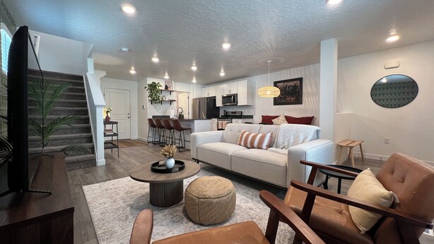 A modern living room with a sofa, ottoman, and chairs, a dining area with a table and chairs, and a kitchen in the background.