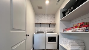 A laundry room with two front-loading washing machines, a shelf with folded towels, and a box of Monopoly on the top shelf.