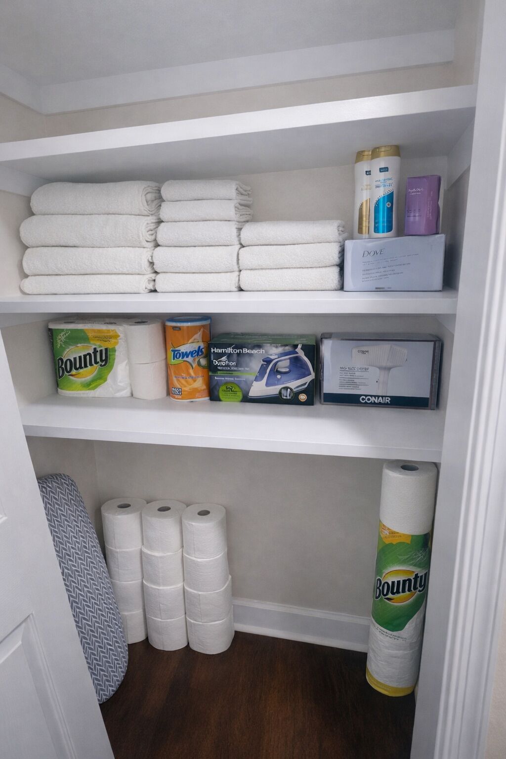 Un armario con toallas blancas dobladas con cuidado, una caja de jabón Dove, una caja de Hamilton Beechnut, una caja de secador Conair y un rollo de papel Bounty.