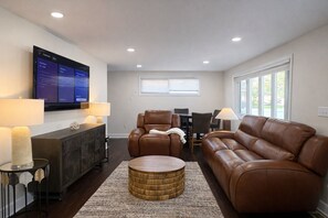 A living room with a flat-screen TV, leather sofas, a round coffee table, and a dining area with a table and chairs.