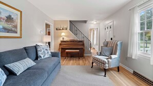 Ein Wohnzimmer mit grauem Sofa, einem gemusterten Sessel, einem Klavier und einer Treppe, die in einen anderen Raum führt.