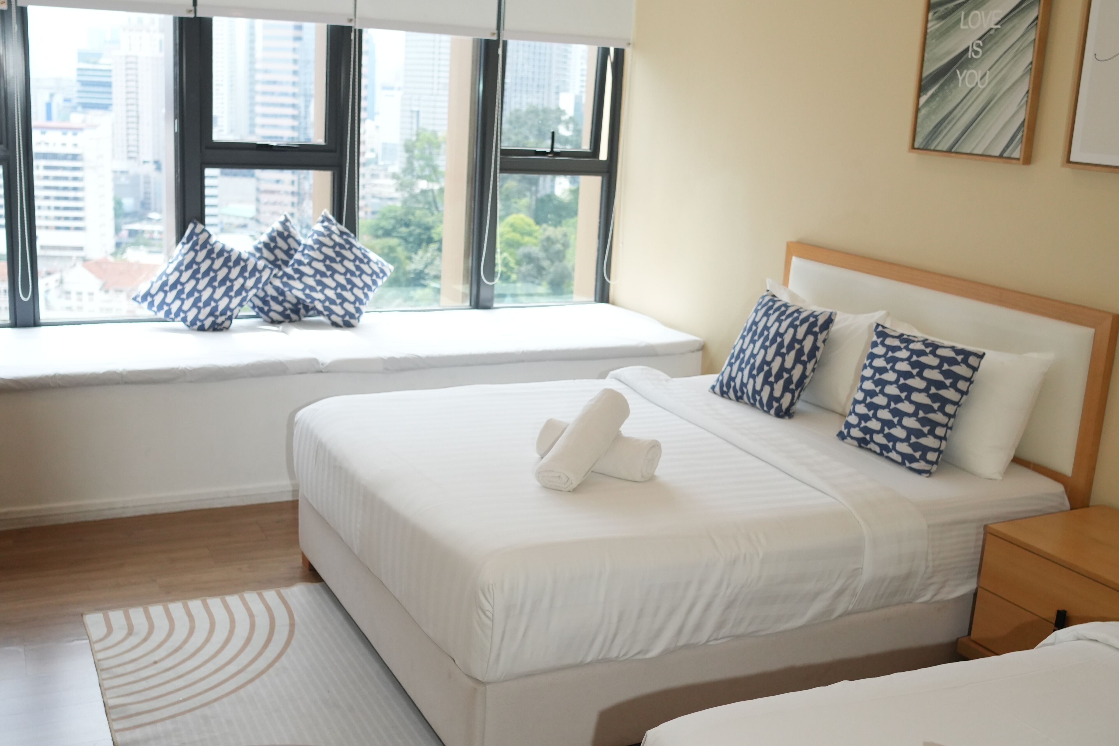 A hotel room with a bed, a window seat with pillows, a bedside table, and a view of the city.