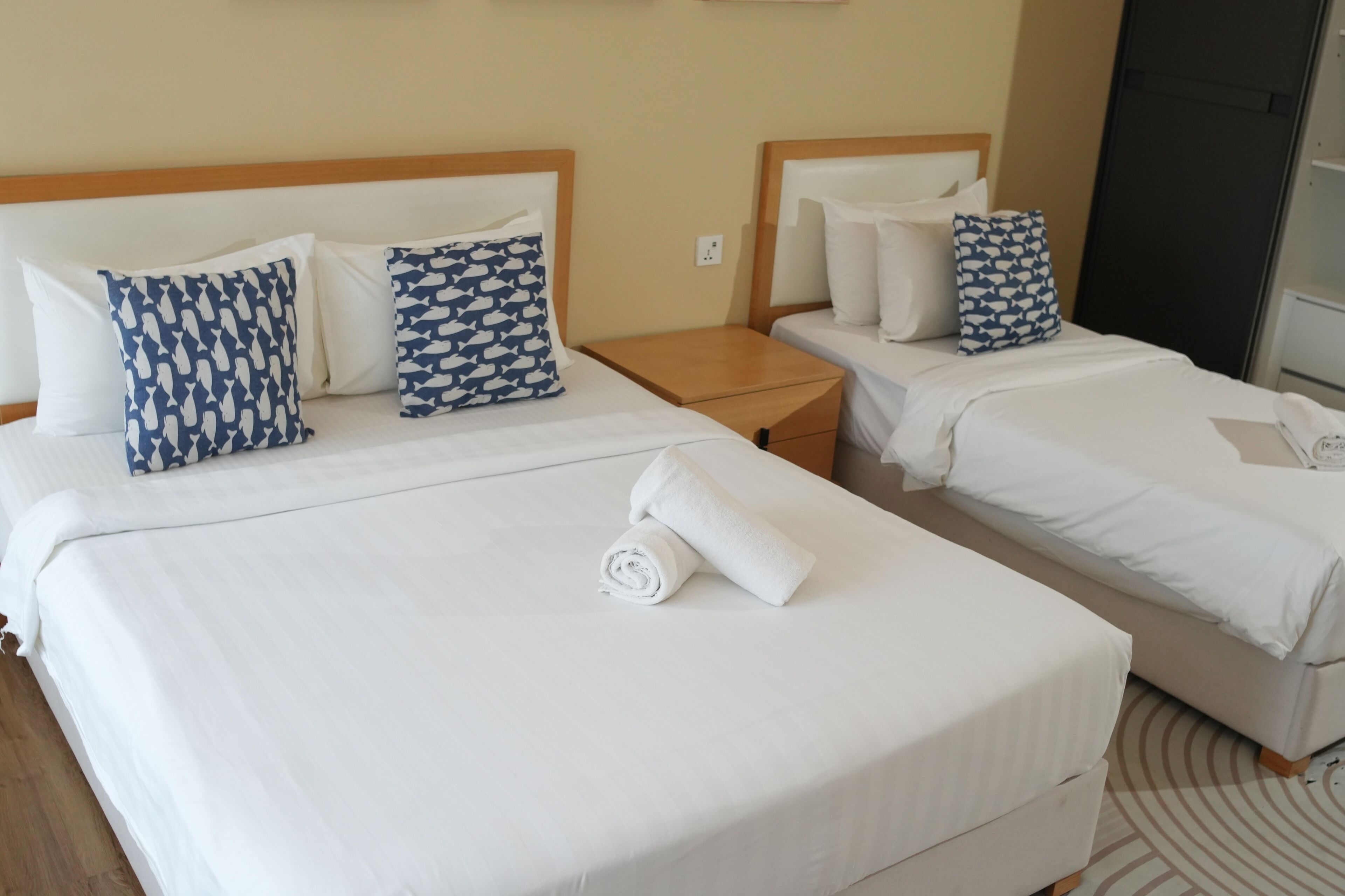 Habitación de hotel con dos camas, cada una con ropa blanca de cama y almohadas estampadas. Hay una mesita de noche de madera entre las camas, y una toalla enrollada sobre la cama.
