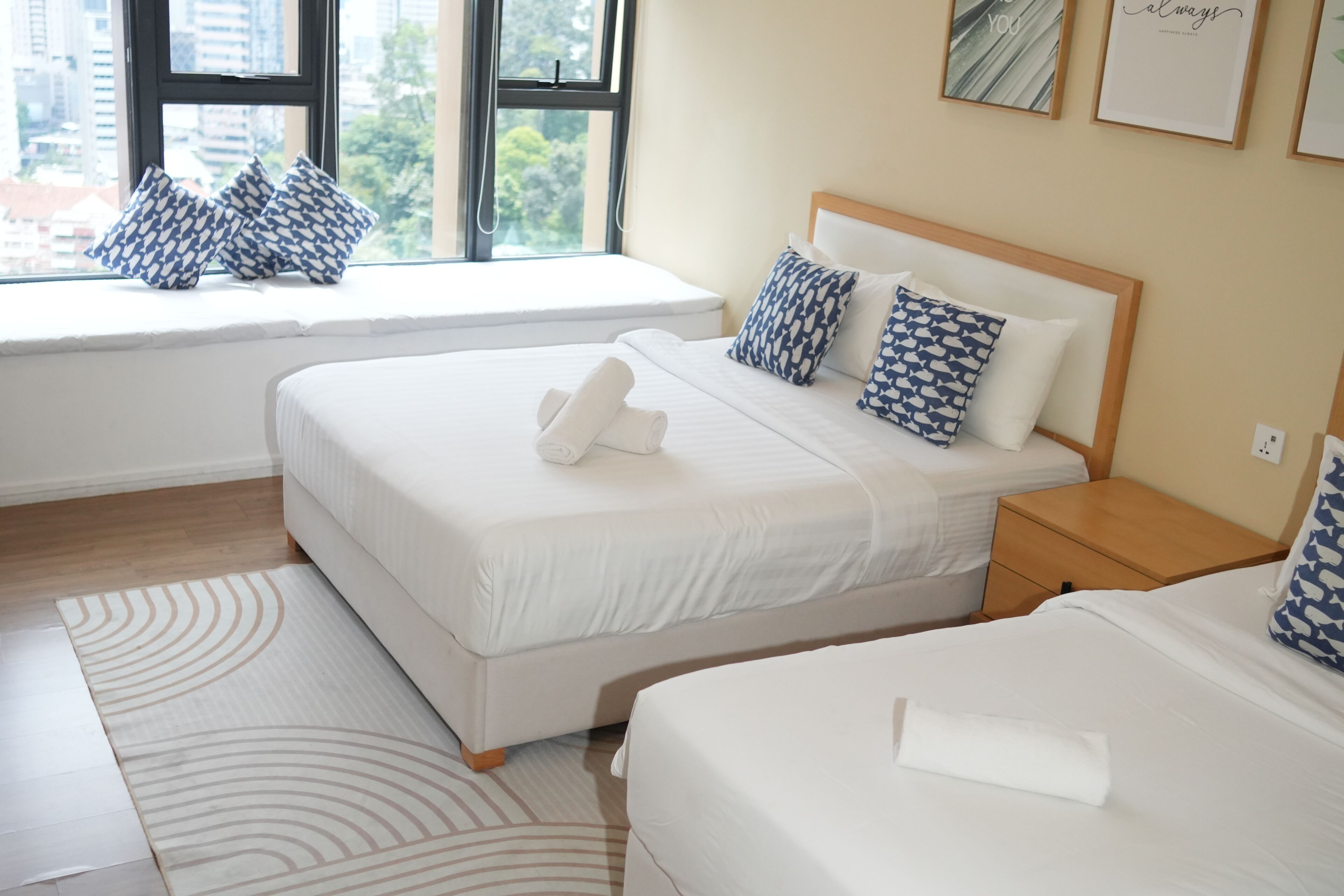 Habitación de hotel con dos camas, un asiento junto a la ventana con cojines estampados, una mesita de noche de madera y vistas a la ciudad.