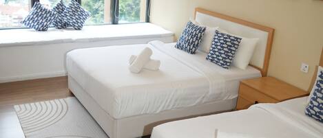 Habitación de hotel con dos camas, un asiento junto a la ventana con cojines estampados, una mesita de noche de madera y vistas a la ciudad.
