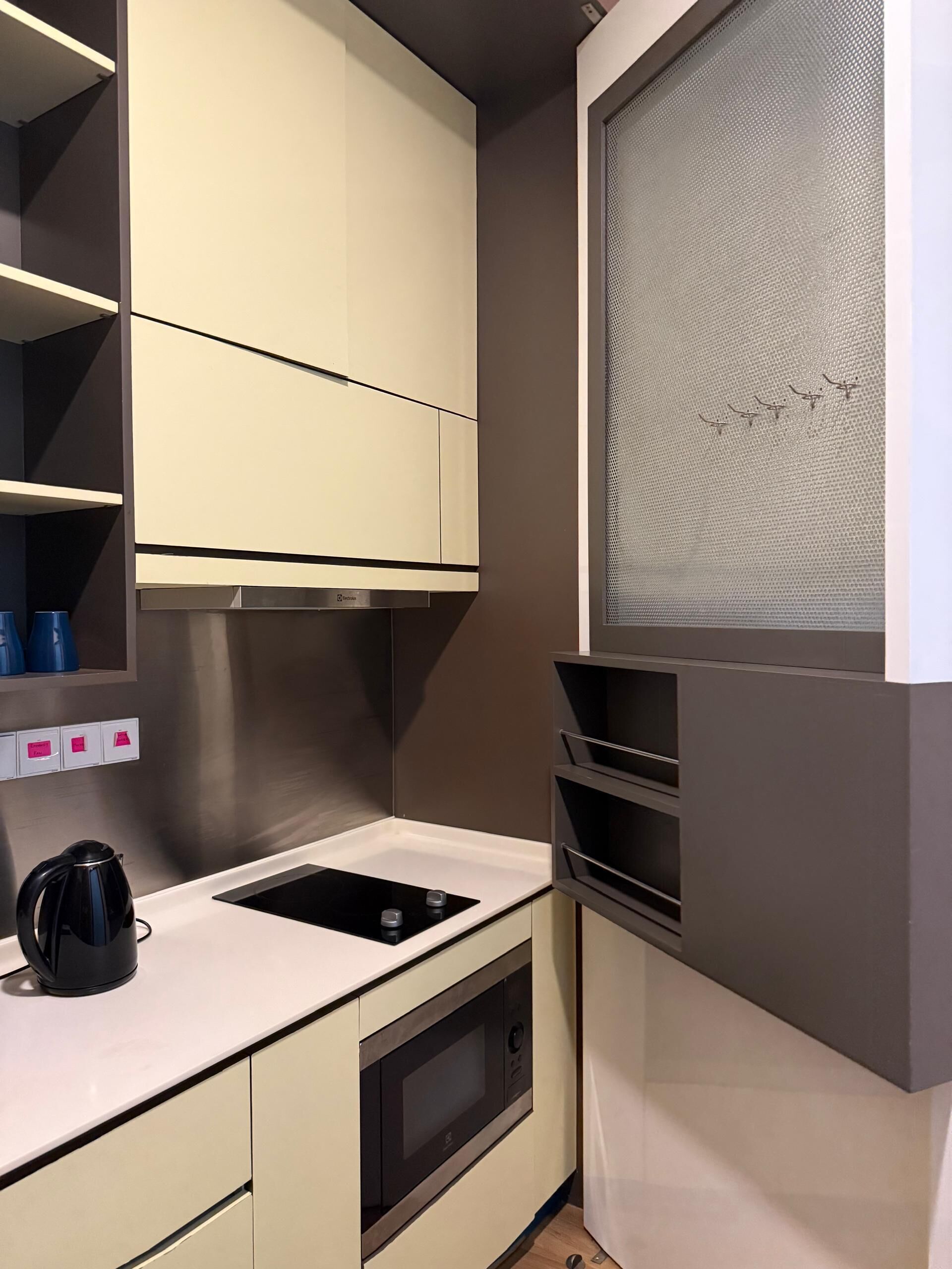 A compact kitchen with a built-in oven, a stovetop, and a microwave. There are cabinets above the counter and a window with a textured blind.