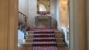 A grand hallway with a marble staircase, a large mirror above a console table, and a red carpet leading to a door.