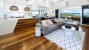 A spacious living area with a wooden floor, a grey sectional sofa, a white coffee table, and a large window offering a view of the outdoors.