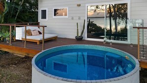 A pool area with a wooden deck, a lounge chair, and a view of the sunset over the water.
