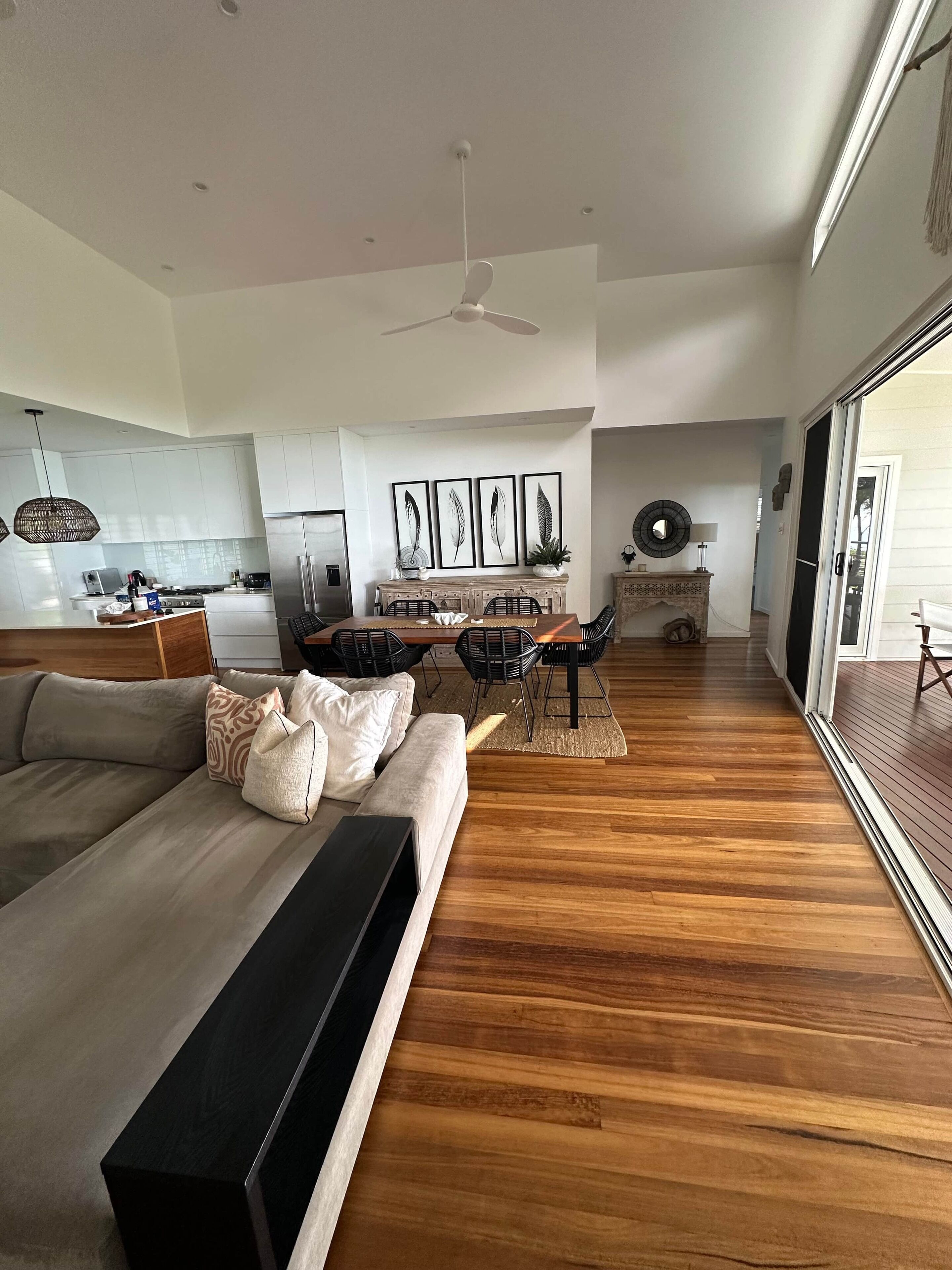 A modern living room with a large sofa, a dining area with a table and chairs, a kitchen in the background, and a large sliding glass door leading to an outdoor area.