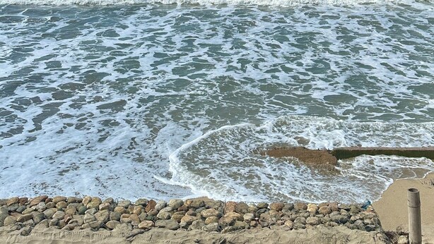 Ein Strand mit Wellen, einer Holzterrasse und einem Gebäude mit einem Reetdach.