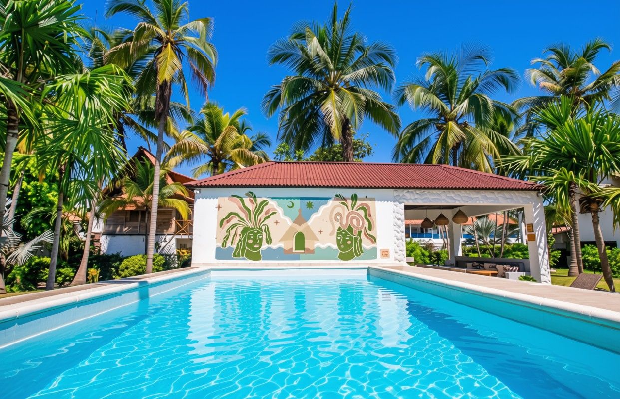 Una piscina con un mural, rodeada de palmeras y un edificio con techo rojo.