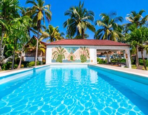 Une piscine à l’eau d’un bleu cristallin, entourée de palmiers et bordée d’un bâtiment orné d’une fresque murale.
