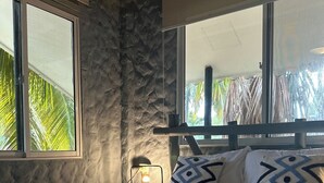 Ein Bett mit weißen Leinentüchern, eine Steinsäule und ein Fenster mit Blick auf Palmen.