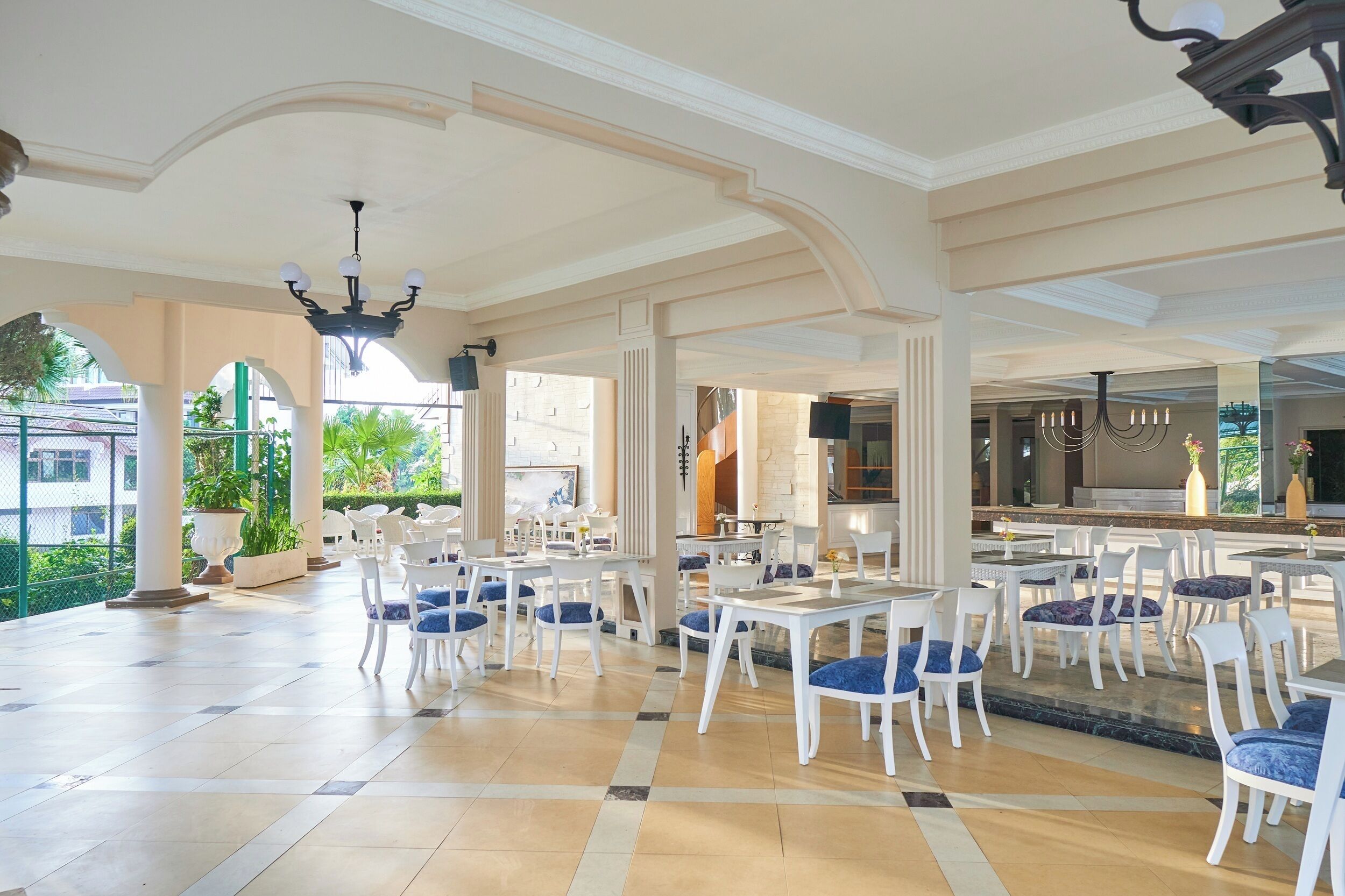 A spacious indoor area with white tables and chairs, a chandelier, and a view of greenery outside.