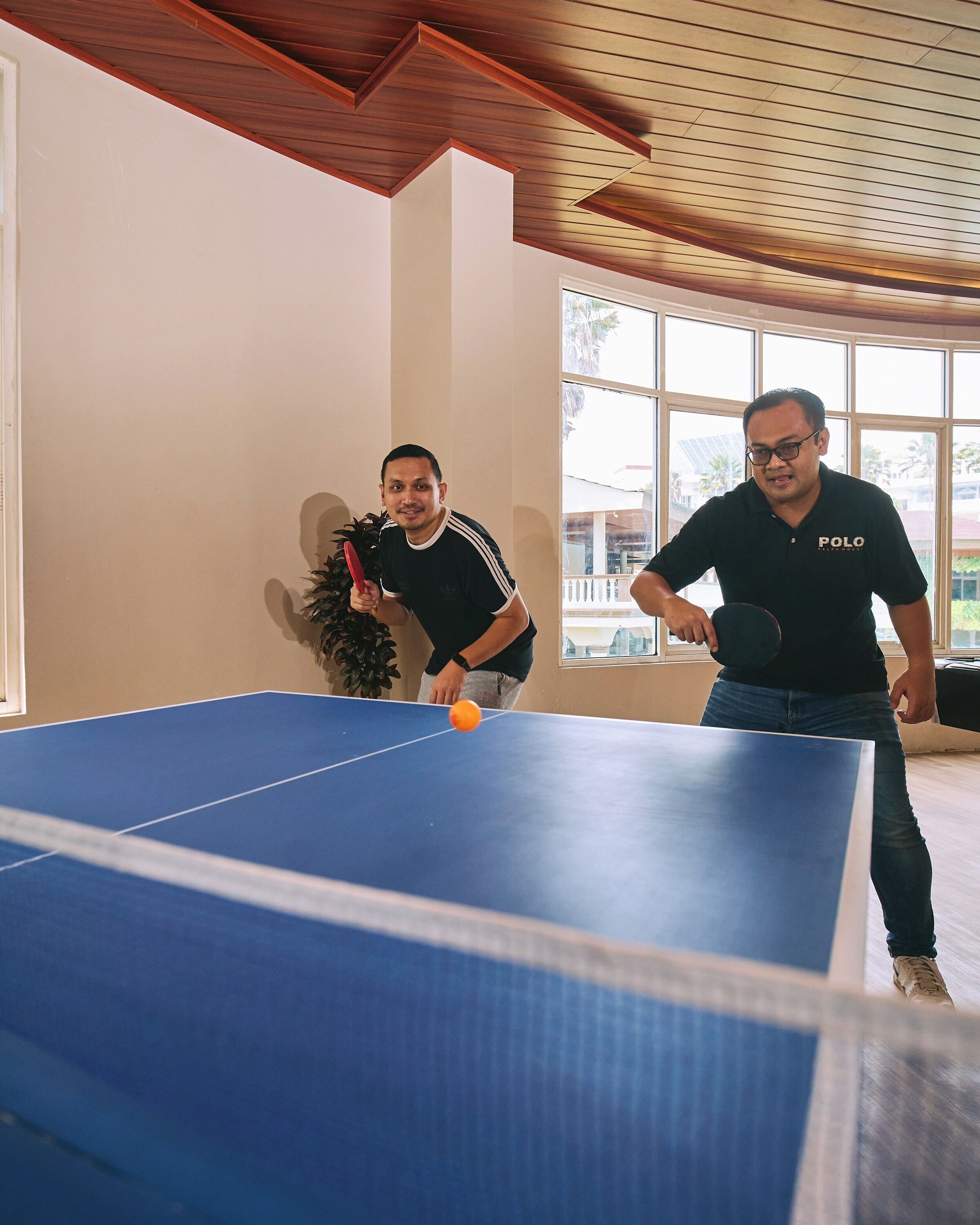 Dos personas jugando ping-pong en una habitación con un ventanal grande y vigas de madera en el techo.