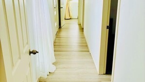 A hallway with wooden flooring and white walls, leading to a room with a window and curtains.