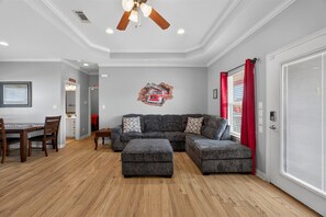 Un salon comprenant un canapé d’angle, un pouf, une table à manger en bois avec des chaises et un ventilateur de plafond.