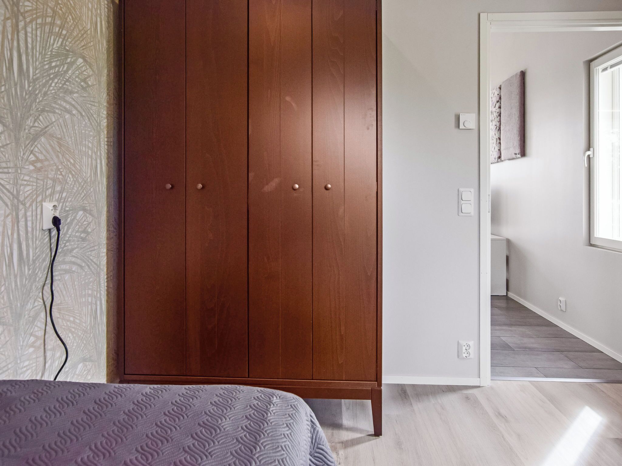 A bedroom with a wooden wardrobe, a patterned bedspread, and a view into another room.
