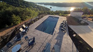 Una piscina con lettini e un tavolo, circondata da un solarium in pietra, con vista su un lago e delle colline sullo sfondo.
