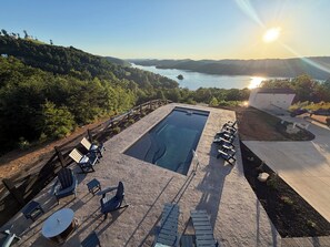 A swimming pool with lounge chairs and a table, surrounded by a stone deck, with a view of a lake and hills in the background.