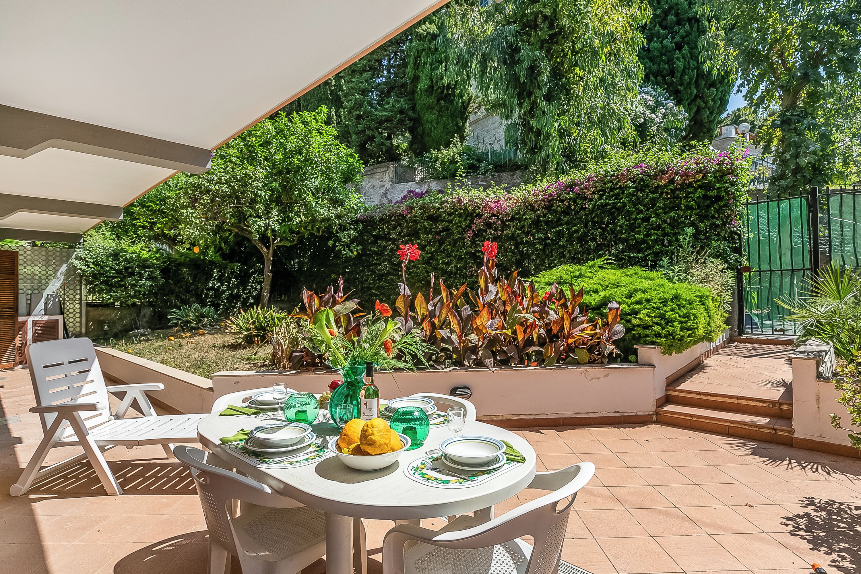 Eine Terrasse mit einem runden Esstisch, umgeben von weißen Stühlen, und ein Garten mit verschiedenen Pflanzen und Blumen im Hintergrund.