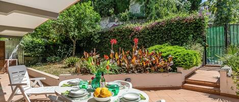Un patio con una mesa redonda lista para comer, rodeada de sillas blancas, y un jardín con diversas plantas y flores al fondo.