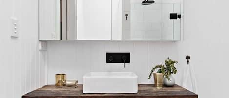 Un baño con tocador blanco, un espejo grande y una ducha visible en el reflejo.