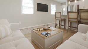 A modern living room with a sofa, a coffee table with decorative items, and a kitchen area with wooden chairs and a refrigerator. - Beautiful Cottage with Water Views - 2 Bedrooms (Delanco)