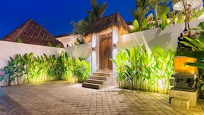A well-lit outdoor area with a thatched-roof structure, steps leading to a door, and lush tropical plants. - Baan Seanok lofts (Pecatu)