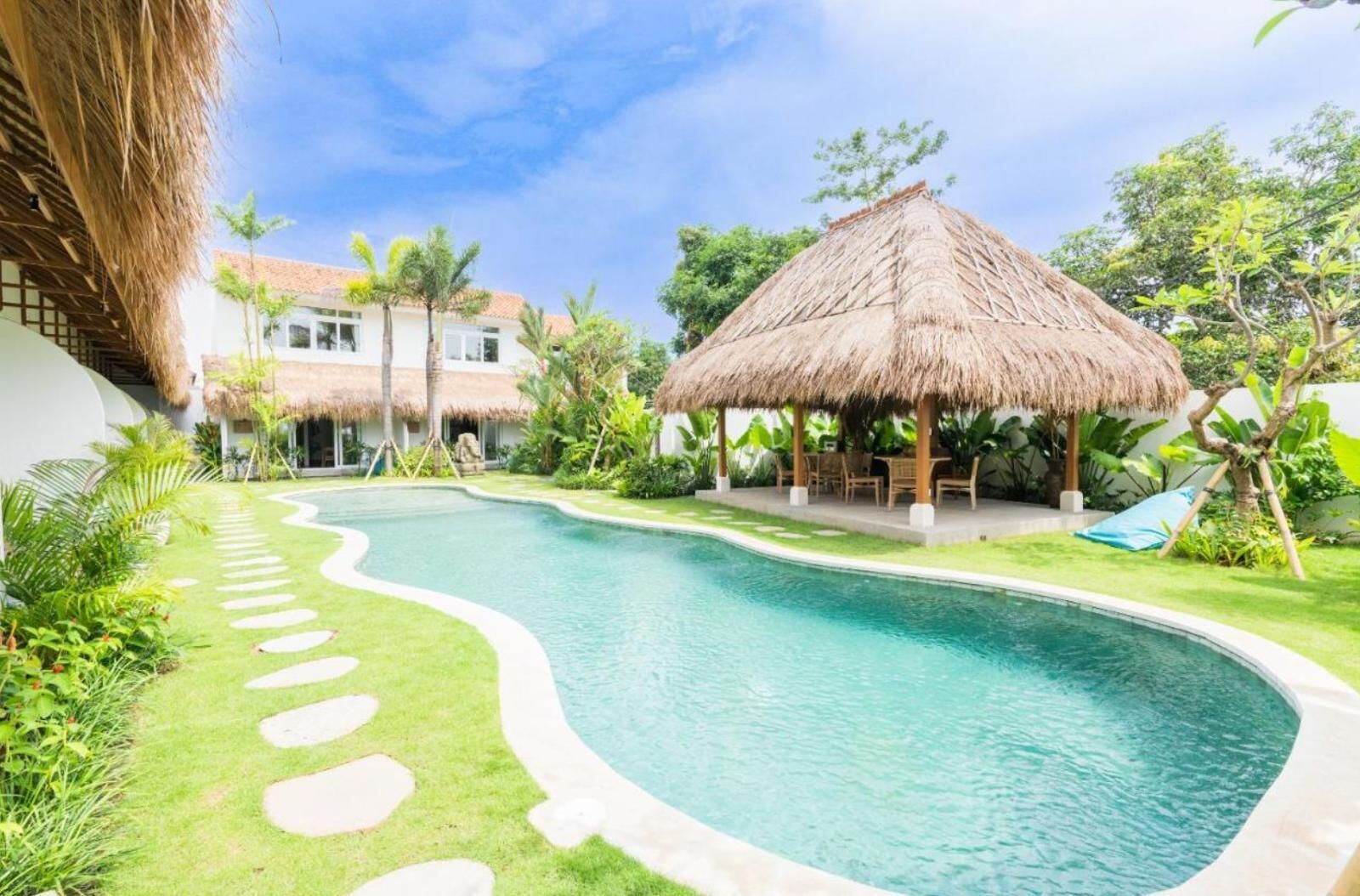 A swimming pool with a thatched-roof structure and lounge chairs, surrounded by greenery and a white building.